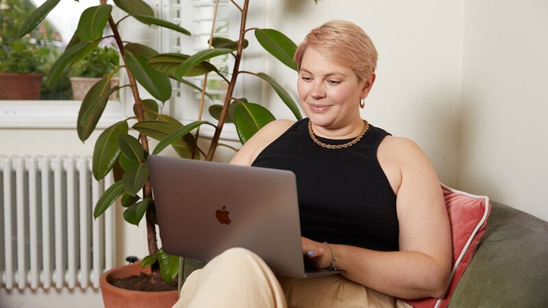 A smiling woman sitting on a couch, using a laptop, learning how to design a product, surrounded by indoor plants.