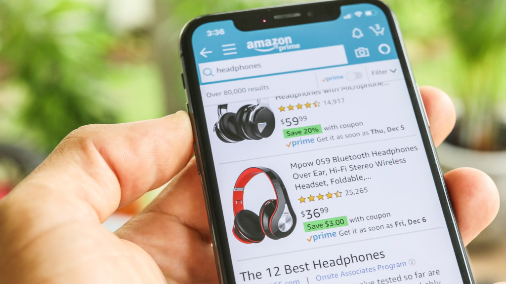 A mobile phone held in one hand with the Amazon app open and various listings of headphones.