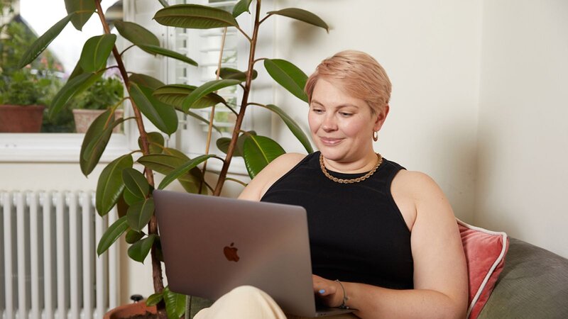 A young woman with short blonde hair, wearing a black shirt is sitting on a couch, working on a laptop with a plant in the background.
