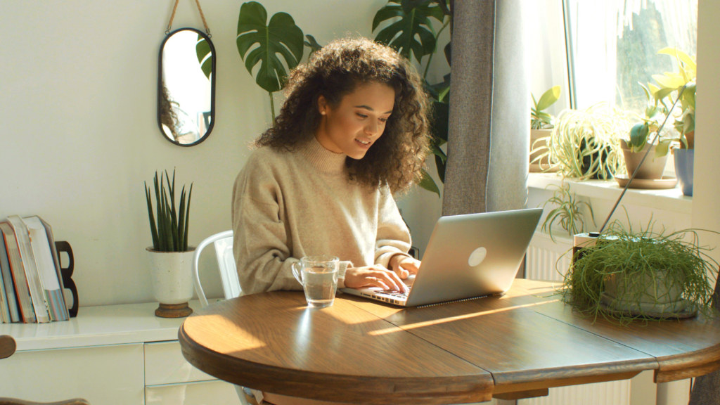 A woman in a gray sweater sitting with a laptop in a kitchen.