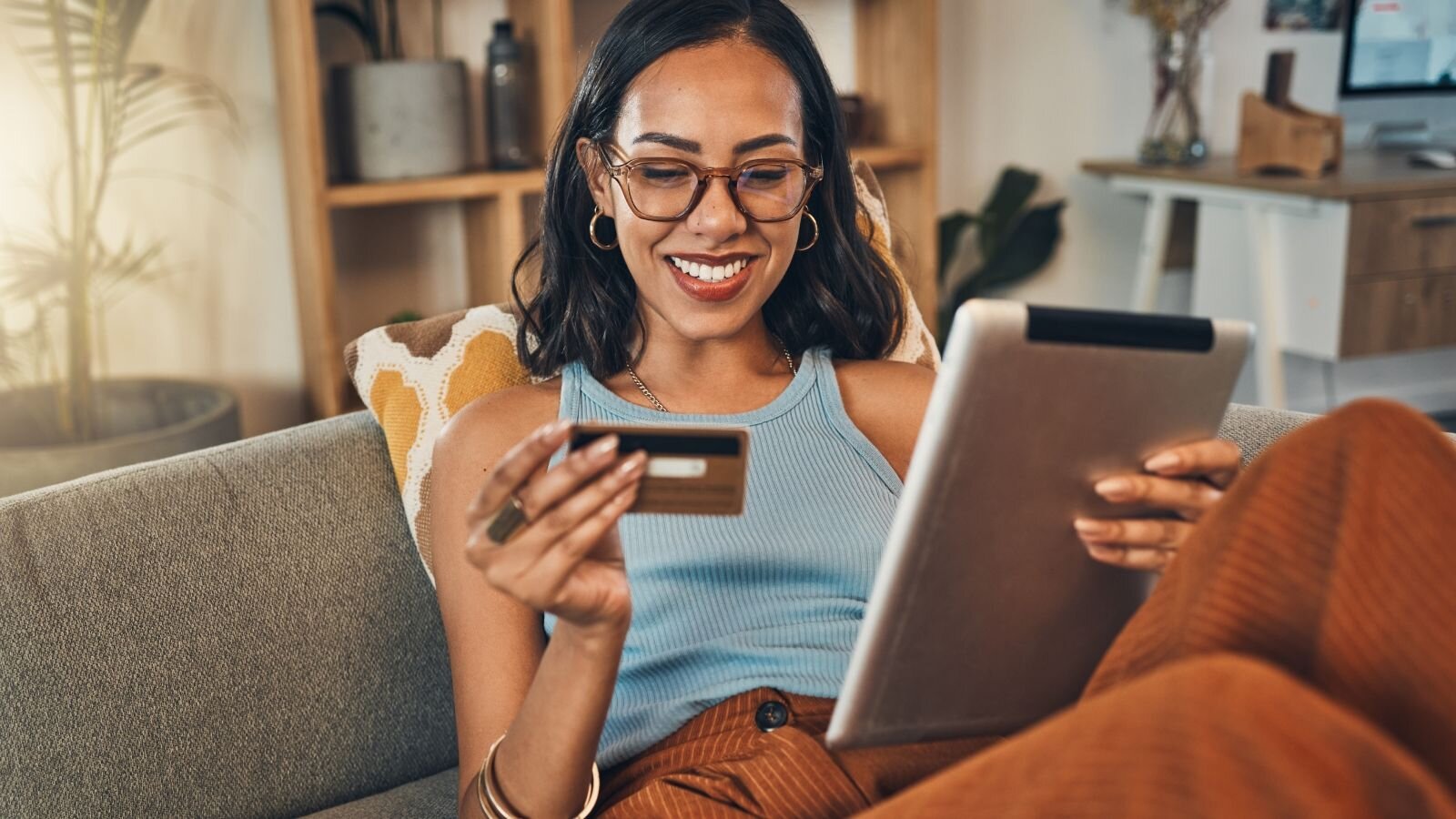 A woman sitting on a sofa, smiling while holding a tablet in one hand and a credit card in the other. She is wearing glasses and has a casual outfit.