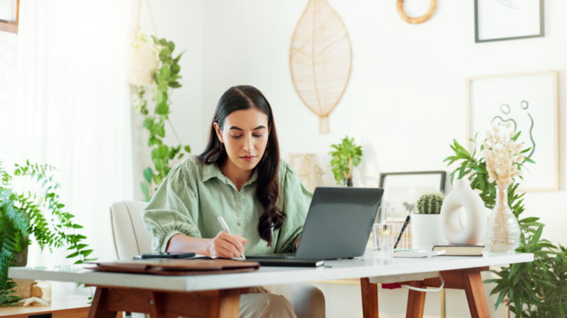 A woman in a green shirt sitting in front of a laptop and writing in a notebook.
