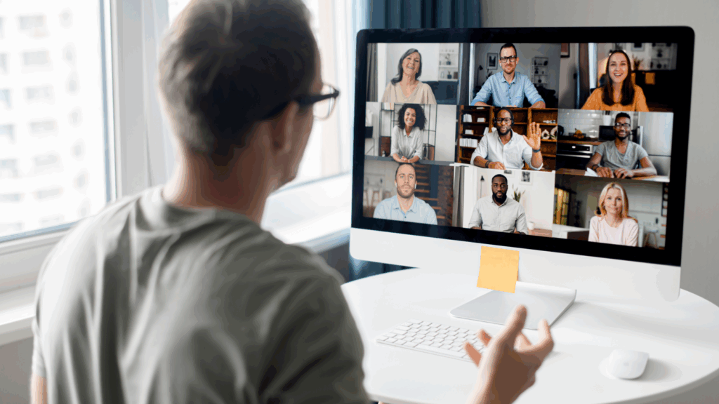 A man sits at a desk, focused on a computer screen displaying a video call with multiple participants.