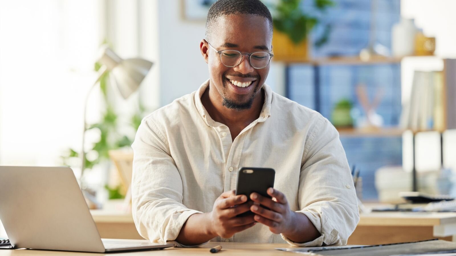 A smiling man in a light shirt is using a smartphone at a desk with a laptop and plants in a bright office environment.