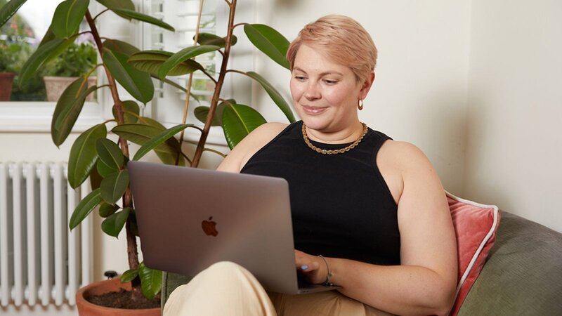 A young, smiling woman in a black shirt sits in a large chair, using a laptop to learn how to create a text design.