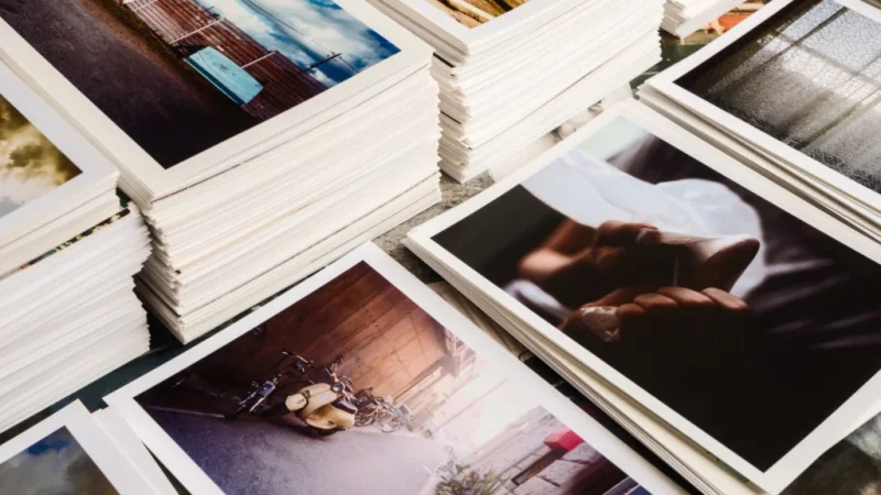 Stacks of glossy photographs on a table, showing various scenes like a vintage motorcycle, serene landscapes, and intimate moments, conveying nostalgia.