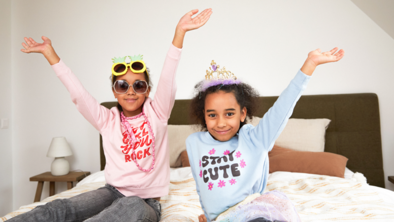 Two young girls are sitting on a bed, wearing colorful outfits and accessories, with their arms raised in a joyful pose.