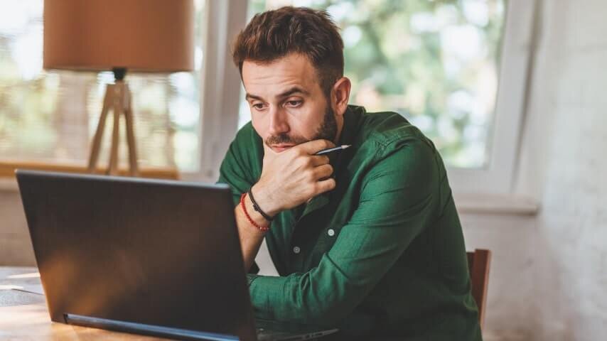 A man wearing a green shirt looking at a laptop screen and thinking.