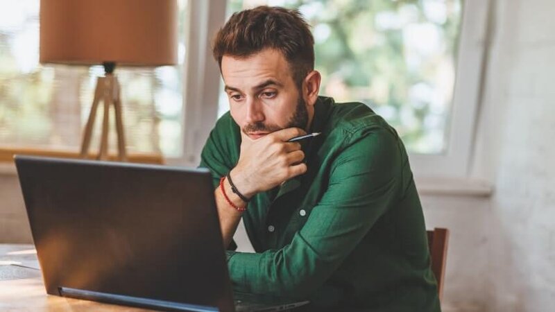 A man wearing a green shirt looking at a laptop screen and thinking.