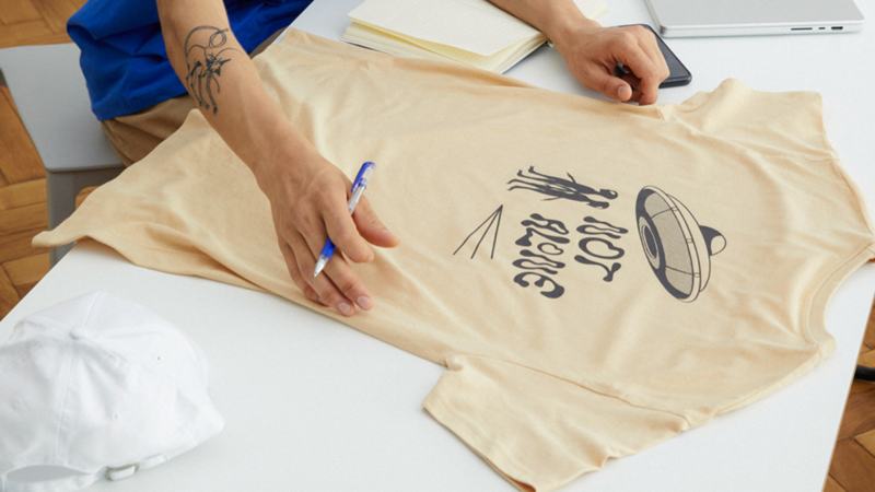 A person’s hands are above a beige alien-themed “Not alone” t-shirt.