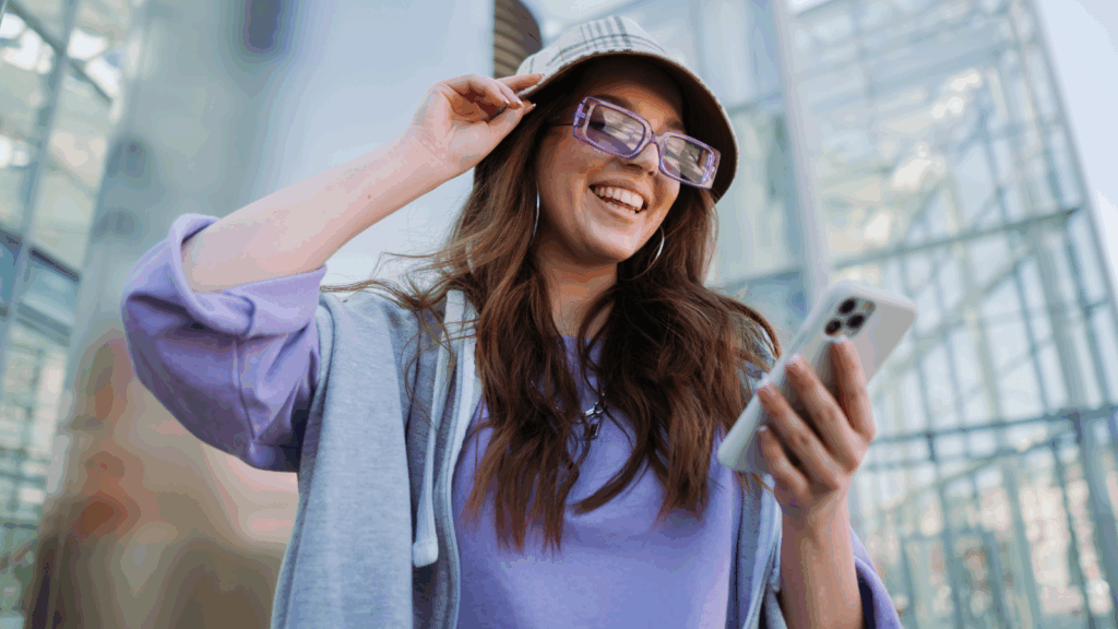 A smiling woman in glasses and a bucket hat is looking at a mobile phone in her hand.