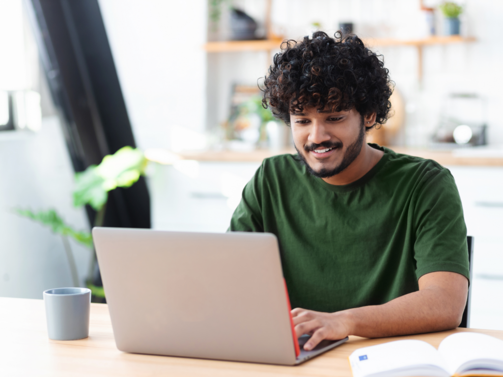 A man with curly hair sits at a table, working on a laptop, brainstorming creative T-shirt marketing strategies.