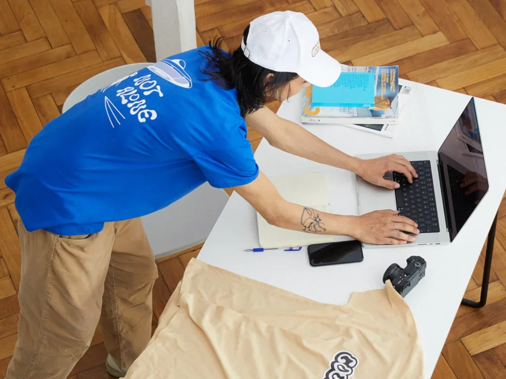 A man wearing a blue "Not Alone" shirt and a white cap, typing on a laptop at a white desk. Nearby are a phone, a camera, and open notebooks.