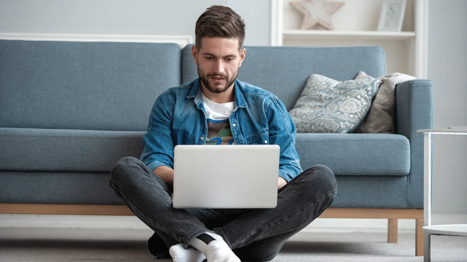 A man sitting on the floor in front of a couch and using a laptop that he has on his lap.