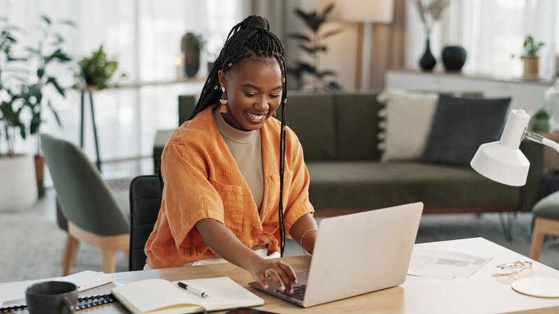 A woman with braided hair is sitting at a desk, smiling as she uses a laptop in a modern, bright interior space.