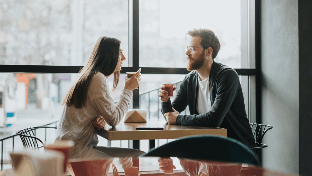 A man and woman seated at a cafe table, engaged in conversation over coffee.