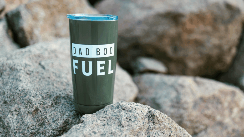 A cup labeled "dad bod fuel" rests on a rock, showcasing its bold design against a natural backdrop.