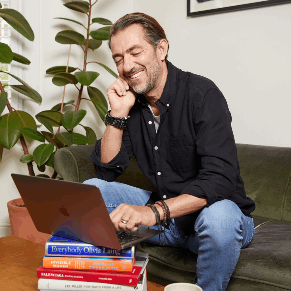 A smiling man using a laptop that’s standing on a stack of books.