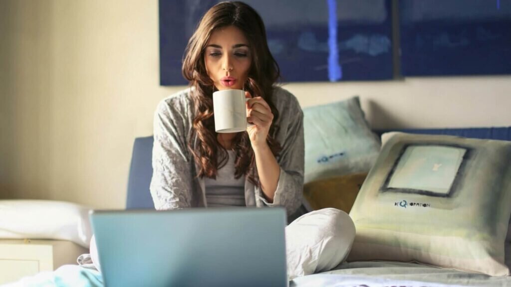 A woman sitting on a bed and drinking coffee, while using a laptop.