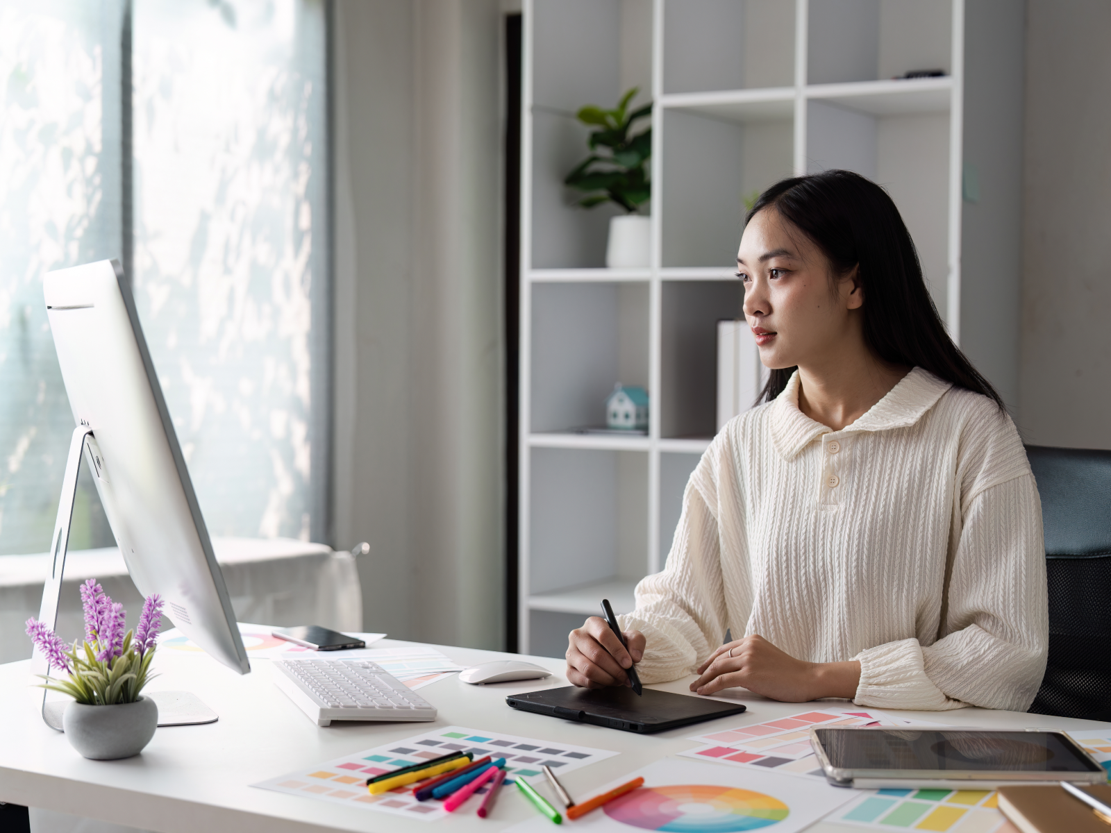 A woman in a white sweater works at a desk using a computer and drawing tablet, preparing her art for printing.
