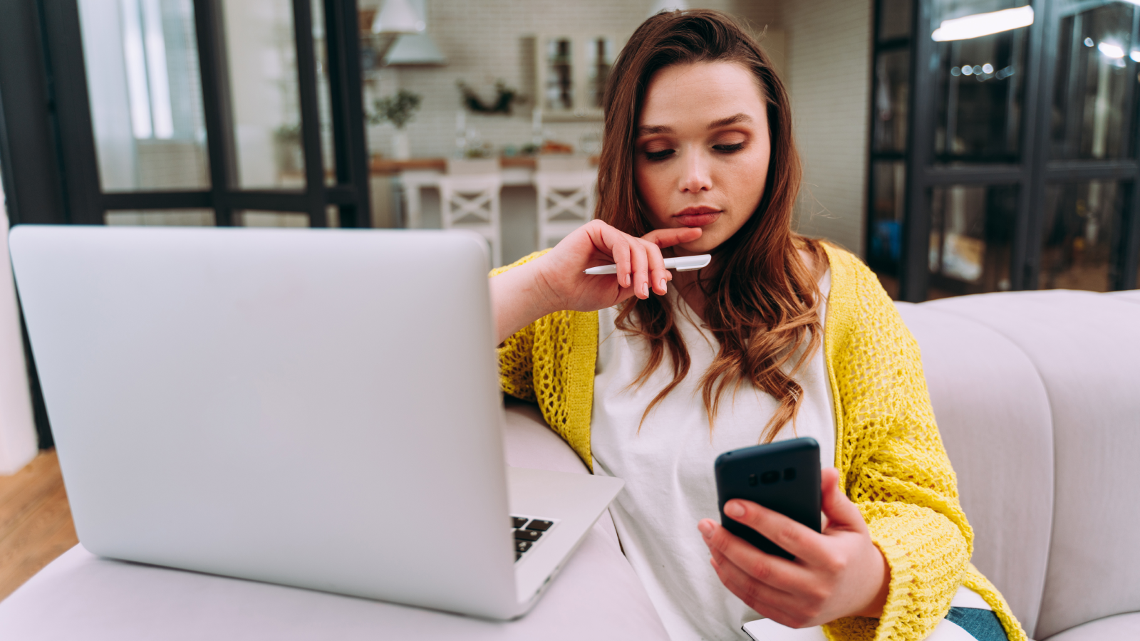 A woman sitting on a couch in front of a laptop, while looking at a mobile phone in her hand.