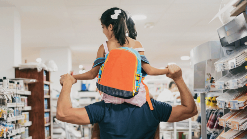 A man carries a child on his shoulders while shopping in a store with a backpack on.