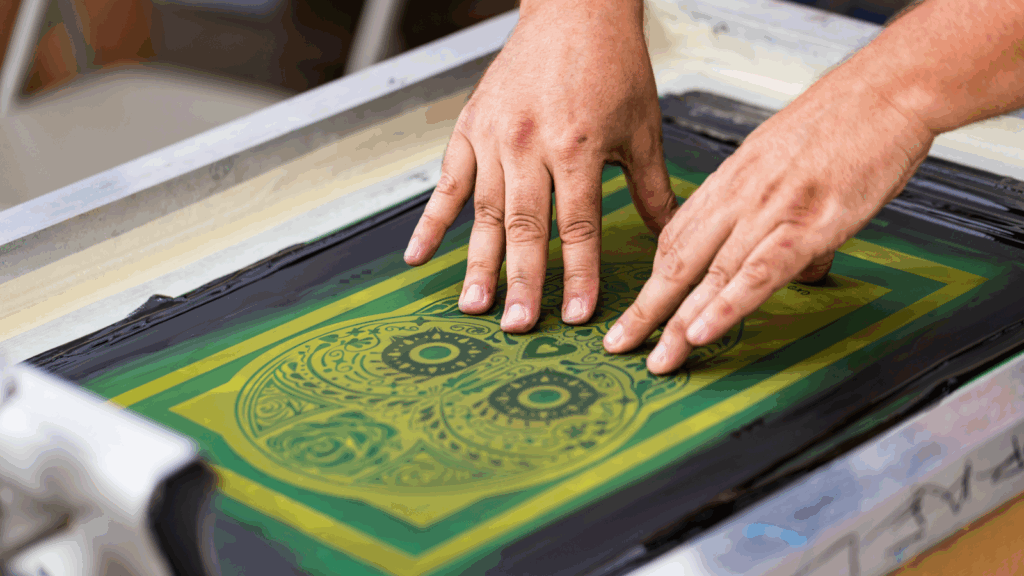A person operates a screen printer to create a design in green and yellow colors.