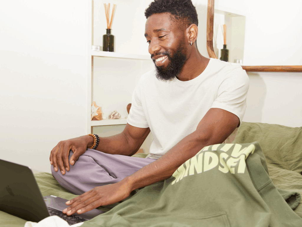 A smiling man sitting on a couch with a laptop on his lap, and a green “Positive mindset” hoodie.