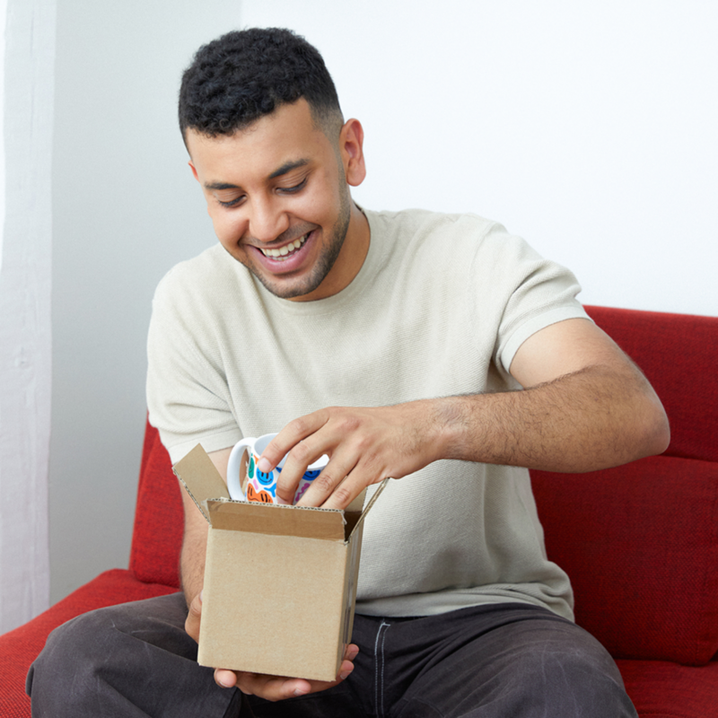 A man sits on a red sofa, smiling joyfully as he opens a cardboard box to reveal a colorful mug inside.
