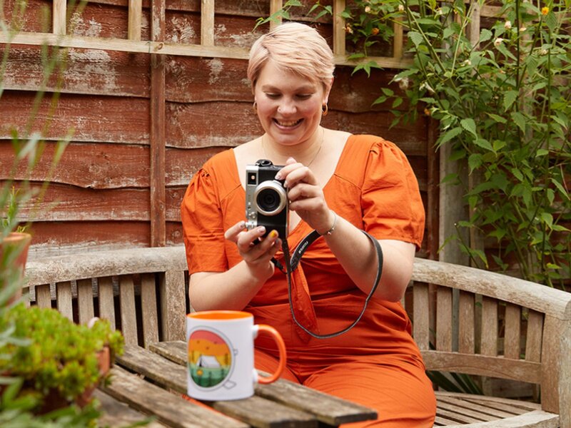 A smiling woman in an orange dress is taking a picture of a colorful mug using a digital camera.