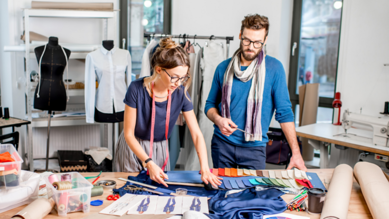 A woman and man work collaboratively in a fashion studio, examining fabric samples and sketches.