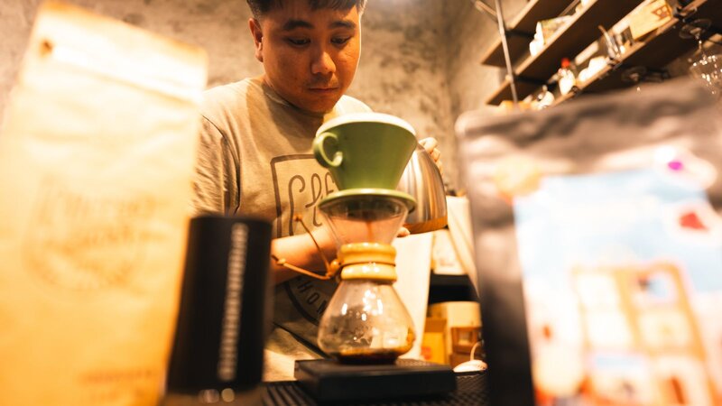 A barista preparing coffee using a siphon brewer, with coffee bags visible in the foreground.