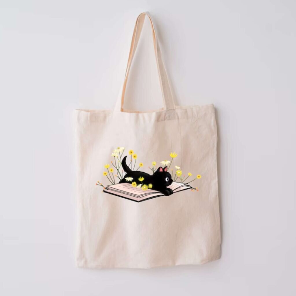 A tote bag with a black cartoon kitten lying on a flowery book.