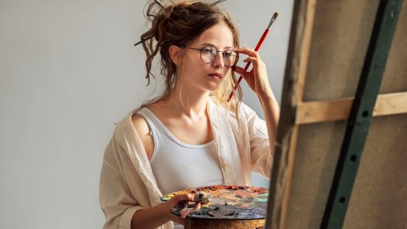 Artist standing in front of her canvas, adjusting her glasses while painting.