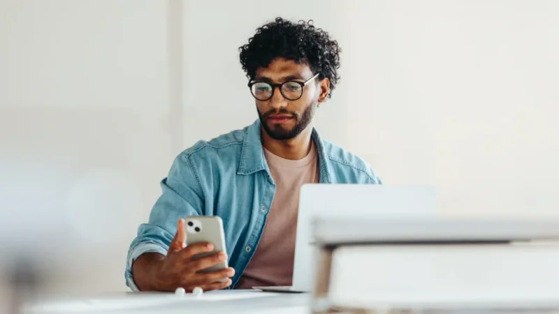 A man with glasses and curly hair looks at his smartphone while sitting at a desk with an open laptop.