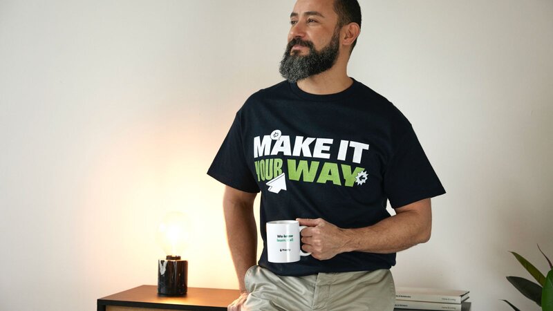 A man with a beard wearing a black t-shirt that says "MAKE IT YOUR WAY" stands beside a table holding a mug.