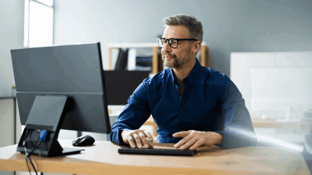 A man in glasses and a blue shirt sits at a desk, working on a computer in an office setting.