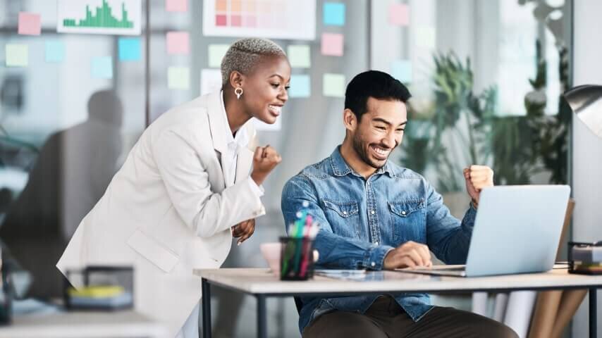 A man and a woman in an office, looking at a laptop screen and smiling.