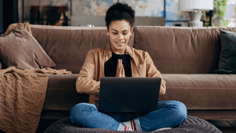 A woman is sitting on the floor with a laptop on her lap, and a couch behind her.