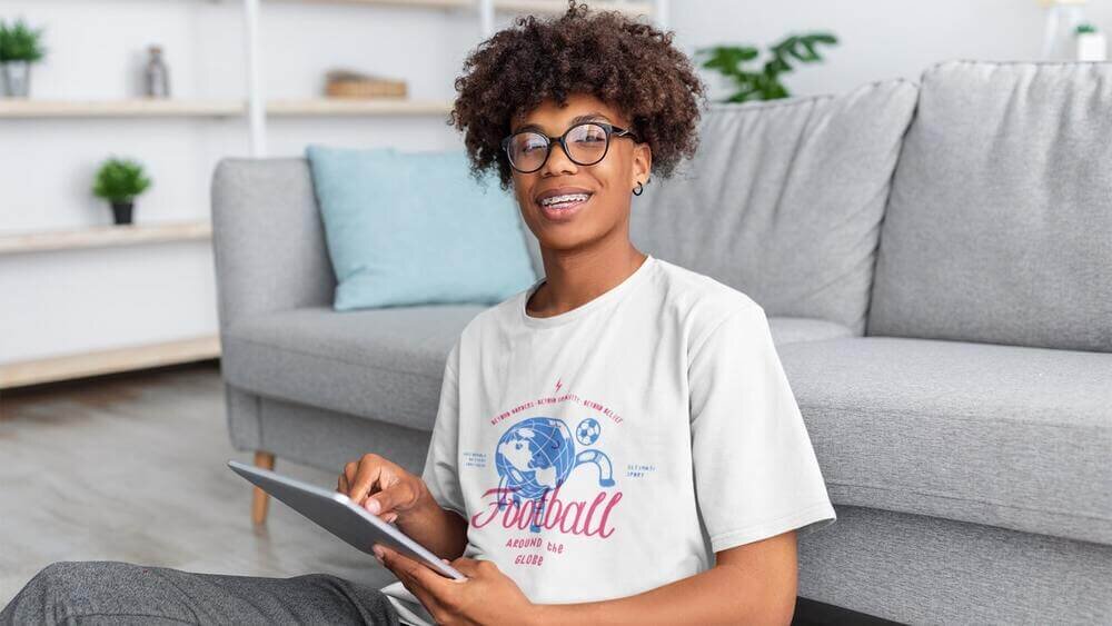 A kid wearing a custom white Gildan 5000B t-shirt with a football print, while holding a tablet.