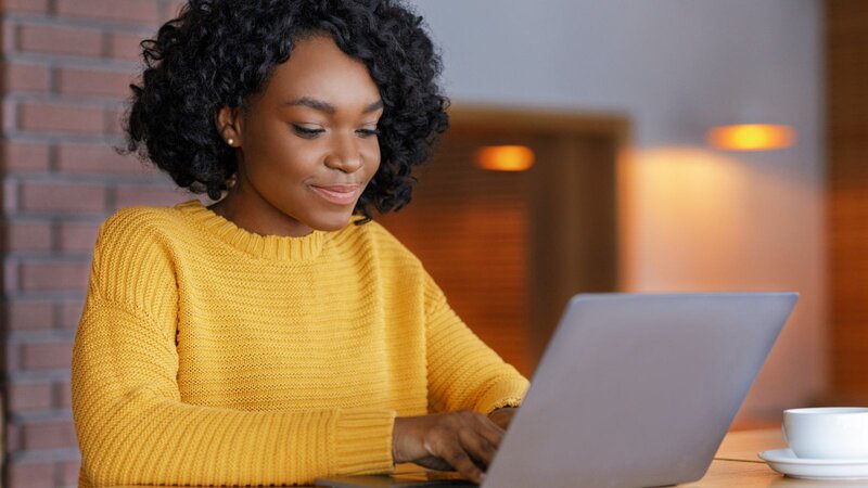 A woman with curly hair in a yellow sweater sitting at a table, using a laptop, learning how to use the Printify API.