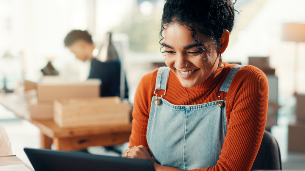 A woman wearing a bright orange sweater and denim overalls smiles at her laptop. The background is softly blurred.