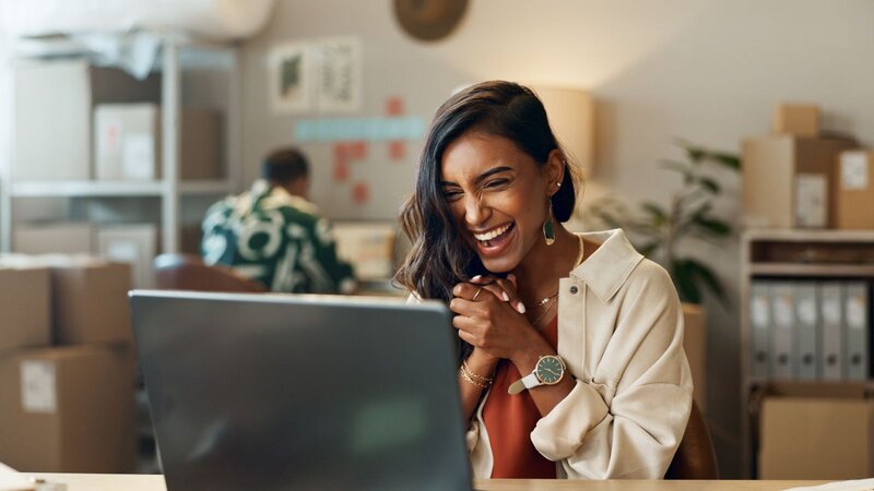 An excited woman clasps her hands and laughs while looking at her laptop in a small business office with shipping boxes and shelves behind her.
