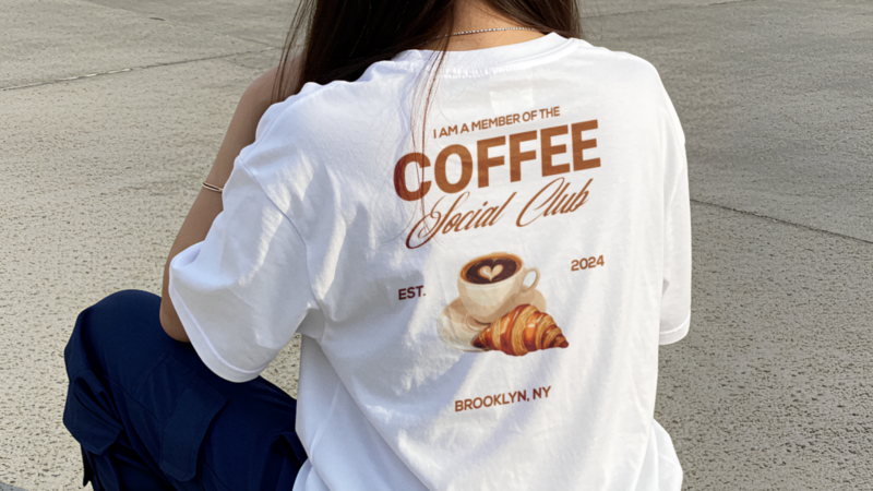 A backshot of a woman wearing a white “I am a member of the Coffee Social Club” with an illustrated coffee cup and a croissant.