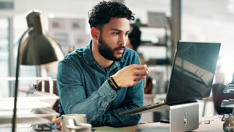 A man with curly hair and a beard is sitting at a desk, focused on his laptop. He is wearing a denim shirt and holding a pen in his right hand.