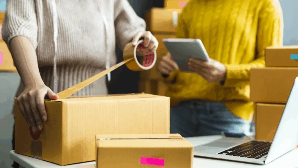 Two individuals collaborate on a laptop surrounded by cardboard boxes in a workspace setting.