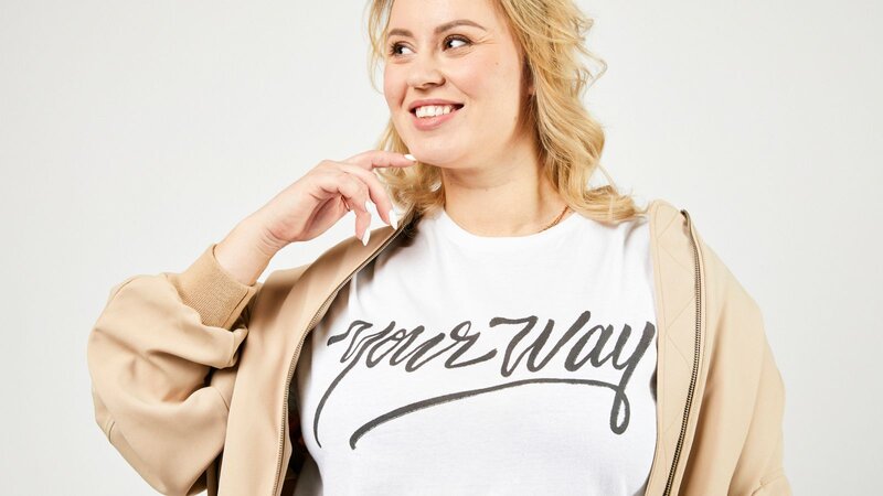 A smiling blonde woman is wearing a white “Your way” t-shirt with a camel jacket on top. White background.