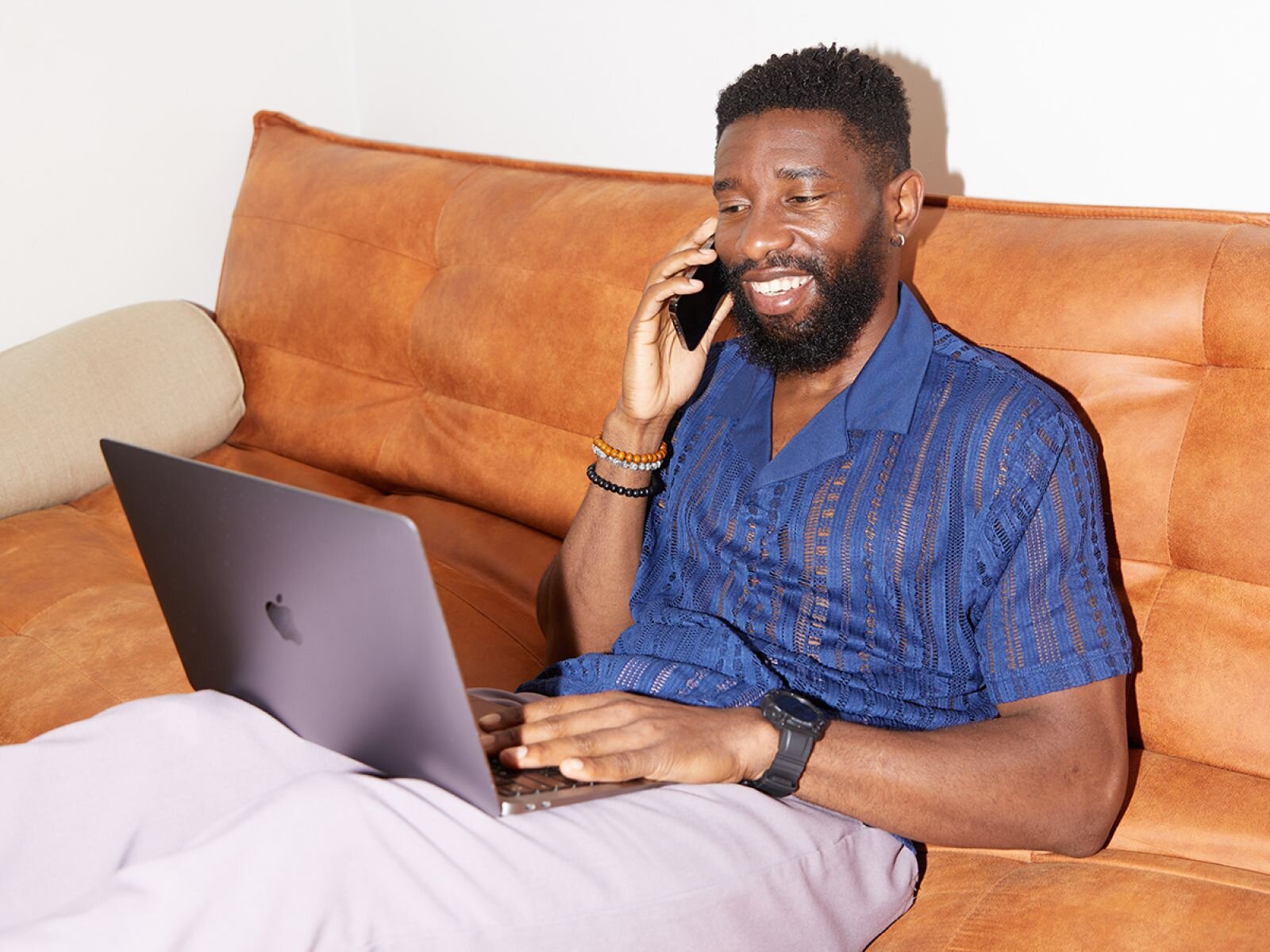 A smiling man in a blue shirt and light pants is sitting on an orange couch with a laptop, learning how to be successful on Etsy.