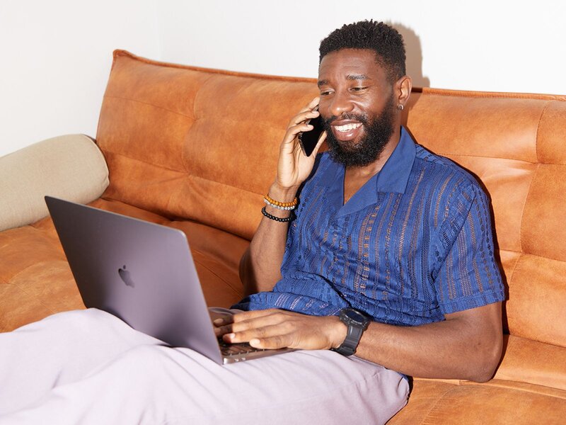 A smiling man in a blue shirt and light pants is sitting on an orange couch with a laptop, learning how to be successful on Etsy.