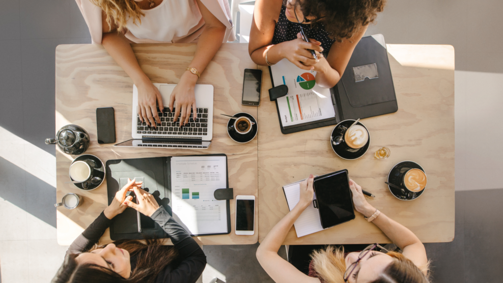 A group of four women collaborating at a table, each using a laptop in a well-lit office environment.